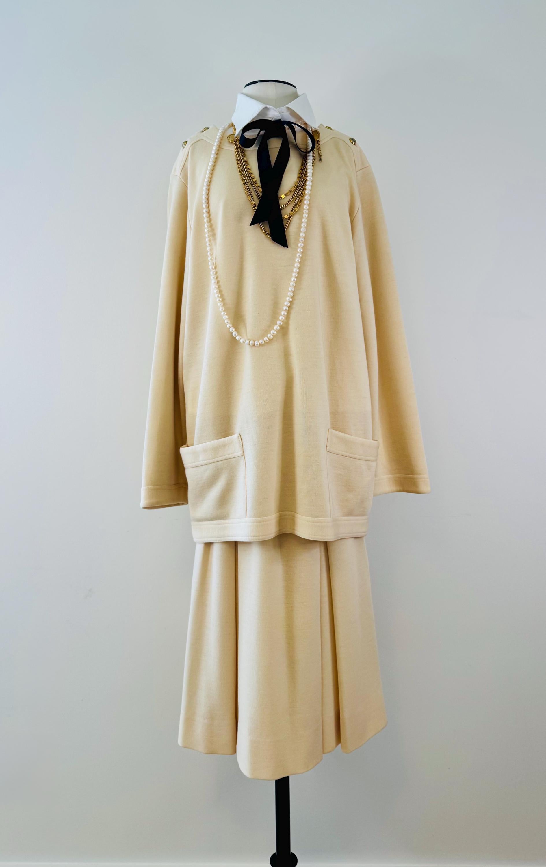 May include: A cream colored sweater and skirt set with a black bow tie and a pearl necklace. The sweater has two pockets and long sleeves. The skirt is a-line and has a pleated design.