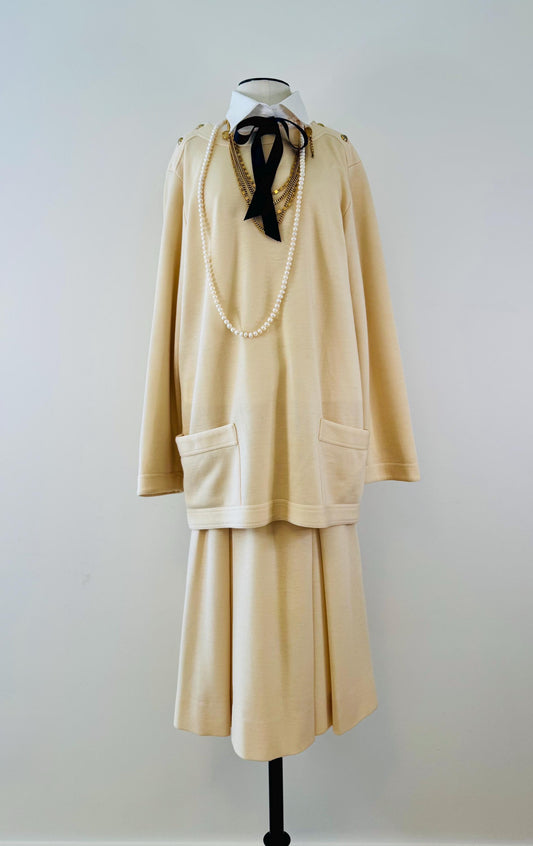 May include: A cream colored sweater and skirt set with a black bow tie and a pearl necklace. The sweater has two pockets and long sleeves. The skirt is a-line and has a pleated design.