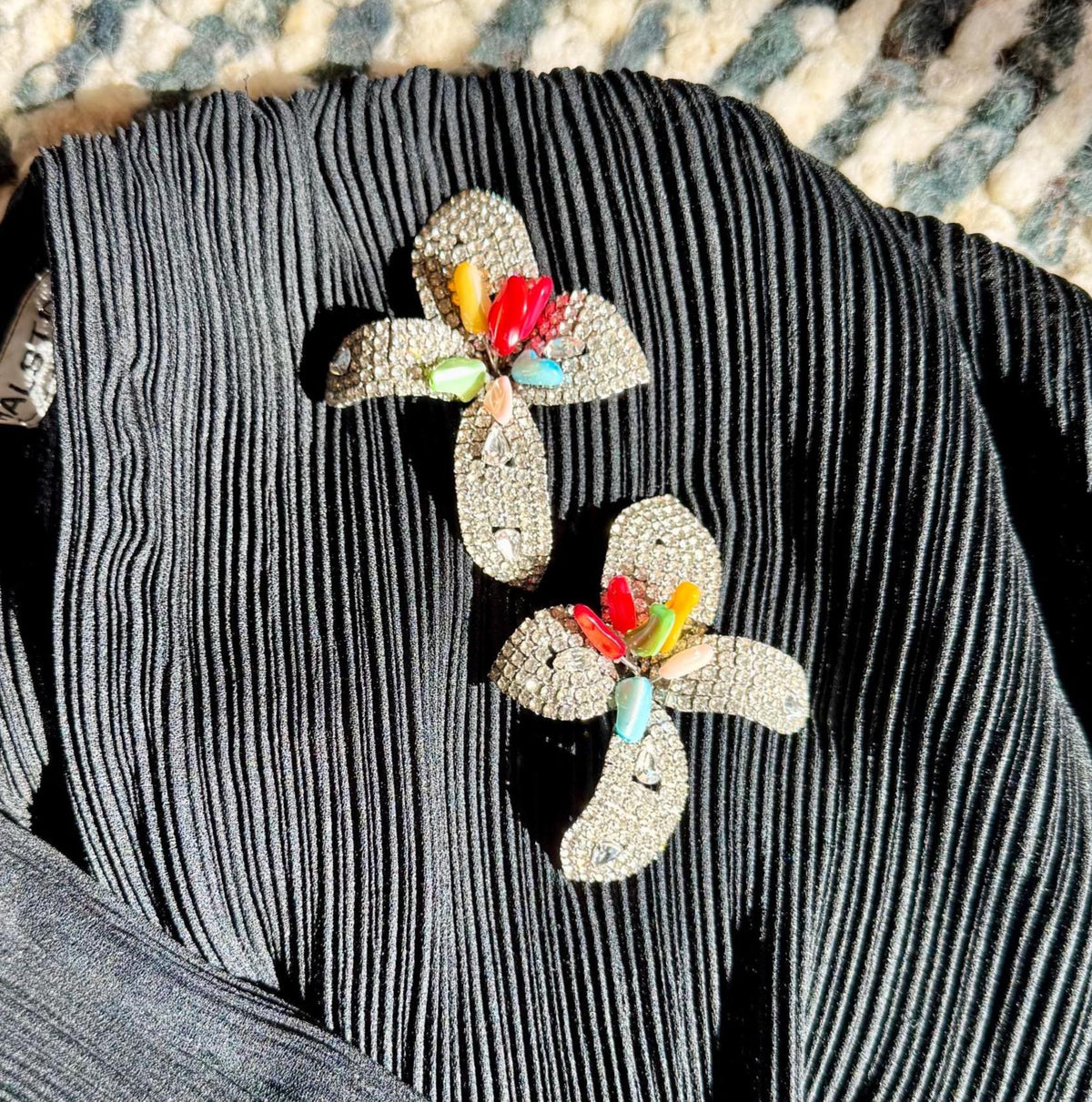 May include: Two flower-shaped brooches with silver-colored rhinestones and colorful gemstone centers. The brooches are set against a dark, vertically-pleated fabric. The gemstones are red, yellow, green, and blue.